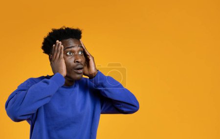 Foto de Impresionado joven guapo emocional negro en suéter azul mirando el espacio de copia para la publicidad, tocando su cara, mostrando asombro en el fondo del estudio amarillo, banner web - Imagen libre de derechos