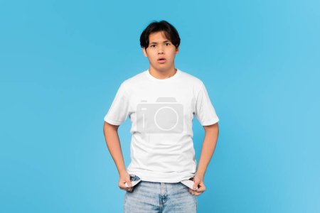 Photo for No Money. Unhappy Japanese Teen Boy Showing Empty Pockets Having Financial Problems Looking At Camera In Shock Posing Standing Over Blue Studio Background. Crisis Concept - Royalty Free Image