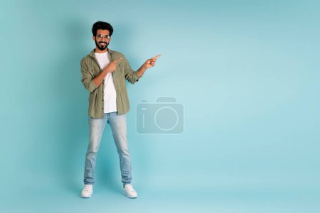 Photo for Happy cheerful handsome young indian man wearing eyeglasses and casual outfit pointing at copy space for advertisement on blue studio background, full length, panorama - Royalty Free Image