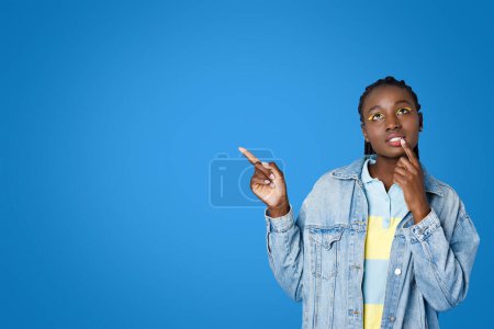 Photo for Exciting offer. Pensive dreamy beautiful young stylish black woman with braided hair and bright yellow eyeshadows hipster touching lip and looking up, pointing at copy space, blue studio background - Royalty Free Image