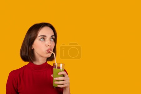 Téléchargez les photos : Jolie brune mince belle jeune femme en verre rouge tenant avec smoothie vert, boire une boisson saine et regarder l'espace de copie. Isolé sur fond de studio orange. Désintoxication, alimentation saine - en image libre de droit