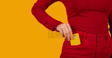 Photo for Close up cropped photo shot of female hand arm putting credit bank card in red jeans pants denim front pocket isolated on orange studio background, copy space. Money finance currency concept - Royalty Free Image
