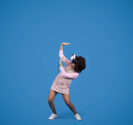 Photo for Emotions from virtual reality. Shocked black little girl in VR glasses plays online game, isolated on blue background, studio. Gadget for fun, childhood, ad and offer, full length - Royalty Free Image