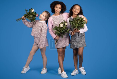 Téléchargez les photos : Joyeuses petites copines internationales avec des bouquets de fleurs profiter du printemps isolé sur fond bleu, studio. Fête, félicitations, émotions de l'enfance et des enfants - en image libre de droit