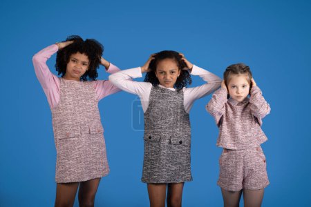 Photo for Sad international little girls in casual covers ears, suffer from noise isolated on blue background, studio. Problems with education, children emotions, stress and bad mood - Royalty Free Image