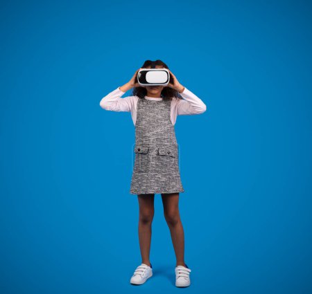 Téléchargez les photos : Souriant petite fille noire en lunettes VR joue jeu en ligne, s'amuse isolé sur fond bleu, studio. Annonce et offre, étude avec dispositif, technologie moderne, émotions de l'enfance et des enfants - en image libre de droit