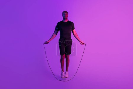 Foto de Deportista negro haciendo ejercicio con cuerda de salto haciendo ejercicio físico posando sobre fondo de estudio de neón púrpura. tiro de longitud completa de hombre deportivo entrenamiento haciendo saltos mirando a la cámara - Imagen libre de derechos