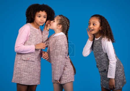 Photo for Shocked curiously funny multiethnic little girls friends in casual whispering secrets and gossip, isolated on blue background, studio. Children emotions, sharing dreams, communication - Royalty Free Image