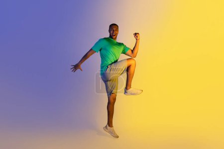 Téléchargez les photos : Entraînement. Happy Sporty Black Guy faisant de l'exercice coude à genou Crunch debout sur fond bleu et jaune néon, souriant à la caméra. Toute la longueur de joyeux sportif motivé. Studio Shot - en image libre de droit