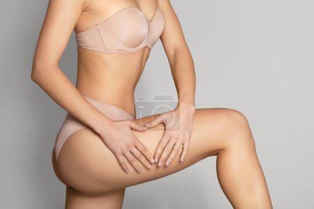 Photo for Love yourself. Cropped view of slim lady in nude panties making heart sign gesture with hands on her thigh, posing over grey studio background. Woman doing anti cellulite massage - Royalty Free Image