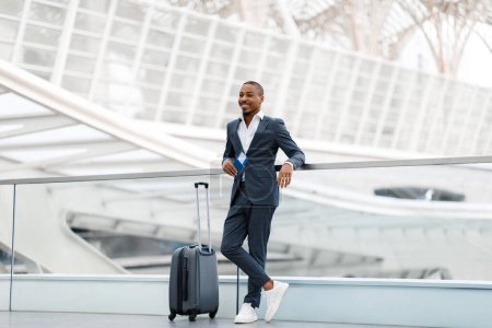 Téléchargez les photos : Bel homme d'affaires noir tenant un passeport avec des billets et debout avec une valise dans l'aérogare, jeune homme afro-américain en costume attendant son vol, profiter d'un voyage d'affaires, copier l'espace - en image libre de droit