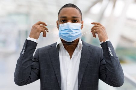 Photo for Portrait Of Young African American Businessman Taking Off Medical Face Mask, Millennial Black Male In Suit Celebrating End Of Coronavirus Pandemic, Enjoying Travelling Without Restriction - Royalty Free Image
