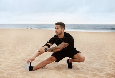 Photo for Young sporty man in sportswear doing leg stretching, preparing for exercising on ocean beach in the morning. Outdoor workout, body care and health care - Royalty Free Image