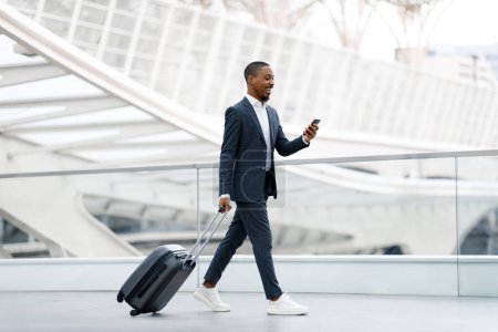 Photo for Handsome black businessman walking with suitcase at airport and using smartphone, young african american male wearing suit browsing internet on cellphone or messaging while going to flight boarding - Royalty Free Image