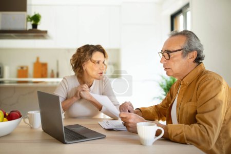 Photo for Concerned senior spouses man and woman sitting at desk in front of laptop, checking bills, making countings, working on family budget together, kitchen interior - Royalty Free Image