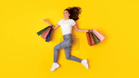 Photo for Shopping And Sales. Cheerful Lady Buyer Jumping Carrying Many Paper Shopper Bags Posing In Studio Over Yellow Background. Woman Shopaholic Smiling To Camera Advertising Seasonal Discounts. Panorama - Royalty Free Image