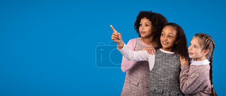 Photo for Shocked smiling diverse little girls in casual clothes have fun, pointing finger and look at empty space isolated on blue background, studio. Surprise, ad and offer for education, children emotions - Royalty Free Image
