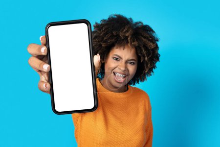 Téléchargez les photos : Émotionnel excité millénaire jolie femme noire bouclée en orange montrant smartphone avec blanc écran blanc maquette sur fond bleu studio, dame africaine montrant affaire en ligne incroyable - en image libre de droit