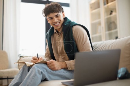 Foto de Sonriente estudiante de Oriente Medio mirando la pantalla de la computadora portátil y tomando notas viendo la conferencia educativa sentada en el sofá en casa. Guy aprendiendo en línea usando la computadora. Enfoque selectivo - Imagen libre de derechos