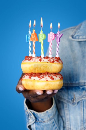 Foto de Recortado de mujer negra en chaqueta de mezclilla sosteniendo pastel dos rosquillas deliciosas dulces uno encima del otro con velas de cumpleaños de colores encendidos, fondo de estudio azul. Concepto de celebración de cumpleaños - Imagen libre de derechos