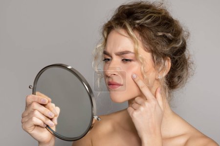 Photo for Upset lady holding magnifying mirror and looking at pimple on cheek, frustrated woman having acne on face, suffering problem skin, standing on grey studio background - Royalty Free Image