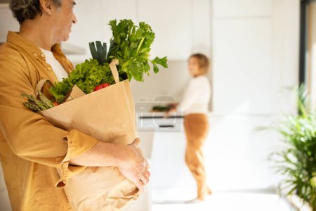 Téléchargez les photos : Homme âgé tenant sac en papier plein de légumes, recevant la livraison du supermarché, debout dans la cuisine, foyer sélectif. Femme préparant le dîner sur fond - en image libre de droit