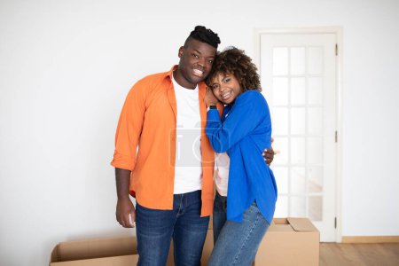 Téléchargez les photos : Portrait d'un couple afro-américain souriant posant parmi des boîtes en carton après avoir déménagé dans une nouvelle maison, joyeux jeunes conjoints noirs embrassant et regardant la caméra, bénéficiant de la réinstallation, Copier l'espace - en image libre de droit