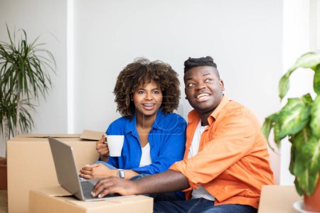 Photo for Happy Black Couple Checking Design Website On Laptop After Moving Home, Smiling African American Spouses Using Computer And Planning Decorations In New Apartment, Sitting Among Cardboard Boxes - Royalty Free Image