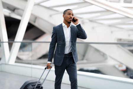 Téléchargez les photos : Homme d'affaires afro-américain parlant sur un téléphone portable tout en marchant avec des bagages à l'aéroport, jeune homme noir en costume ayant conversation de téléphone portable tout en allant à la porte d'envol, copier l'espace - en image libre de droit