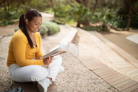 Téléchargez les photos : Concept d'éducation. Joyeux brésilien étudiant fille lecture livre tout en étudiant à l'extérieur, assis dans le parc ou sur le campus de l'université, la préparation pour des conférences ou des cours, espace libre - en image libre de droit