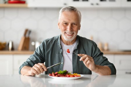 Hombre mayor sonriente guapo comiendo sabrosa comida en la cocina en casa, caballero anciano feliz sentado en la mesa y mirando a la cámara, disfrutando de deliciosa comida, sosteniendo tenedor y cuchillo, espacio de copia