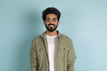 Photo for Happy handsome millennial indian guy in stylish casual outfit and eyeglasses smiling at camera, posing alone on blue studio background, copy space. Millennials lifestyle concept - Royalty Free Image