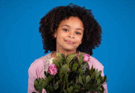 Foto de Positiva niña afroamericana con ramo de flores disfrutar de cumpleaños, aislado sobre fondo azul, estudio, de cerca. Celebración navideña, felicitación y emociones infantiles - Imagen libre de derechos