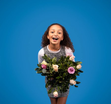 Photo for Laughing african american little girl pupil fresh with bouquet of flowers isolated on blue background, studio. Kid emotions, holiday celebration, congratulation at spring, childhood - Royalty Free Image