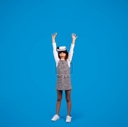 Foto de Inspirado sonriente niño negro en gafas VR juega juego en línea, se divierte, manos arriba aislado sobre fondo azul, estudio, longitud completa. Tecnología, infancia y emociones de la realidad virtual - Imagen libre de derechos