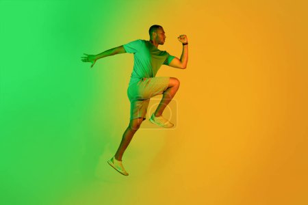 Photo for Determined African American Runner Guy Jumping, Exercising In Green Neon Light Over Yellow Studio Background. Fit Sportsman Running Looking Aside. Side View, Full Length Shot - Royalty Free Image