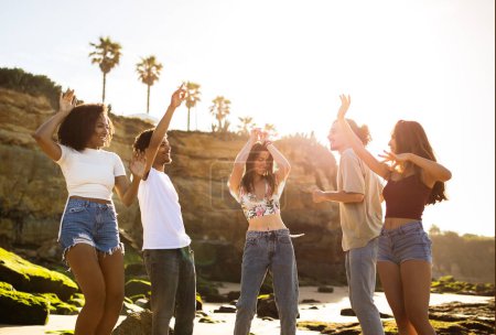 Photo for Glad excited young multiracial people enjoy music, dance, have fun together, enjoy trip on beach. Friendship in summertime, entertainment, holiday vacation and travel party at sunset - Royalty Free Image