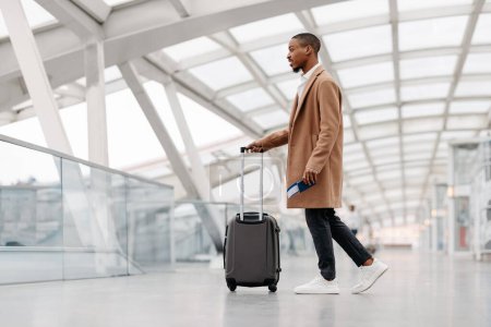 Téléchargez les photos : Beau Noir portant un manteau élégant marchant avec une valise à l'aéroport, Jeune Afro-Américain Homme Passant Terminal Hall, Aller à la porte de départ de vol, Voyager seul en avion, Vue latérale - en image libre de droit