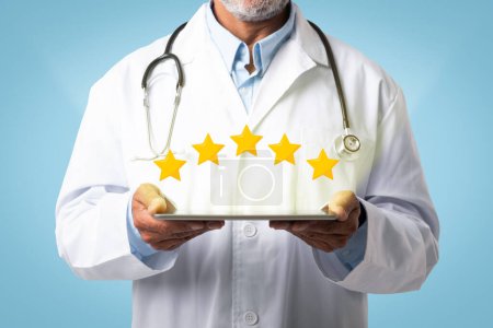 Photo for Senior european doctor in white coat with stethoscope hold tablet with stars on blue studio background. Reviews, rating, device and app for health care, medicine service and consultation - Royalty Free Image