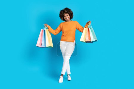 Photo for Season sale, black friday, exciting shopping deal. Happy attractive young african american woman with bushy hair showing purchases colorful shopping bags, isolated on blue studio background - Royalty Free Image