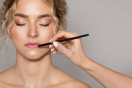 Photo for Professional makeup artist doing nude makeup for caucasian young woman, applying lipstick with brush over grey studio background, closeup, copy space - Royalty Free Image