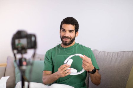 Photo for Cheerful young middle eastern man blogger shows headphones, shooting video on camera in living room interior. Delivery order, blog review, online shopping remotely at home - Royalty Free Image
