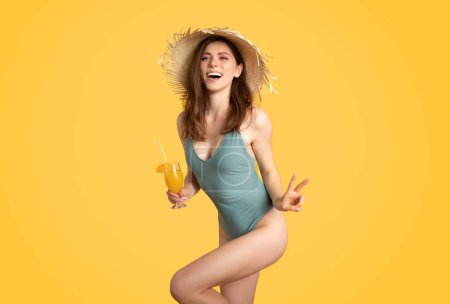 Téléchargez les photos : Joyeuse jeune femme en maillot de bain dégustant un cocktail d'été exotique et montrant un signe de paix, posant et souriant sur fond de studio jaune. Dame excitée s'amuser sur les vacances à la plage - en image libre de droit