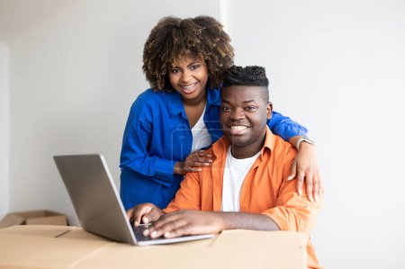 Foto de Pareja de familia negra feliz usando el ordenador portátil juntos mientras se muda a una nueva casa, sonriendo a los cónyuges afroamericanos jóvenes que compran en línea en la computadora, usando cajas de cartón como escritorio, primer plano - Imagen libre de derechos