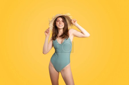 Foto de Retrato de una alegre dama europea posando en traje de baño y con sombrero de paja, de pie sobre fondo amarillo del estudio. Playa vacaciones o piscina concepto de fiesta - Imagen libre de derechos