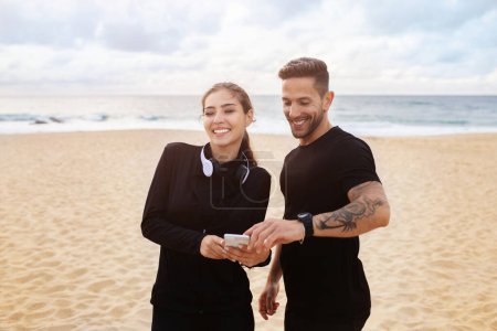 Photo for Happy european man and woman preparing for workout, chatting before morning run on the beach of ocean, lady and guy using smartphones and talking outdoors - Royalty Free Image