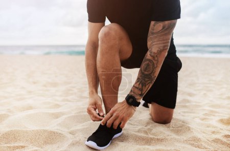 Photo for Man in sportswear tying laces of his shoes while jogging on ocean beach, close up of legs, cropped view. Young male running outdoors, enjoying healthy active lifestyle - Royalty Free Image