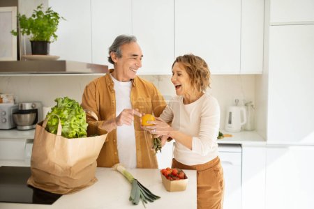 Téléchargez les photos : Joyeux couple de personnes âgées arrivant du supermarché avec sac d'épicerie et déballer dans la cuisine, conjoints âgés profiter de la vie, passer du temps à s'entraider - en image libre de droit