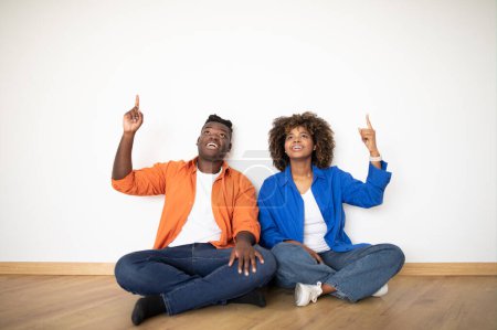 Foto de Pareja negra feliz señalando hacia arriba mientras que se sienta en el piso cerca de la pared vacía en su nuevo apartamento, los cónyuges afroamericanos sonrientes alegres que planean el diseño en casa juntos después de mudarse - Imagen libre de derechos