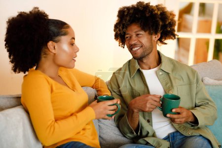 Photo for Cheerful Black Spouses Drinking Coffee Posing With Cups And Talking, Enjoying Domestic Weekend Sitting On Couch At Home. Flirt And Romantic Love Concept. Selective Focus On Man - Royalty Free Image